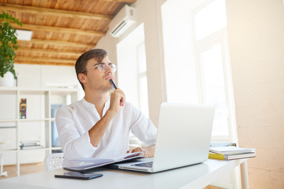 Indoor shot of thoughtful concentrated young businessman wears white shirt and spectacles with laptop and smartphone thinking and writing in notebook at the table in office