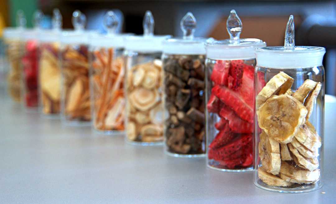 Na białym, błyszczącym blacie stoją szklane pojemniki wypełnione suszonymi owocami