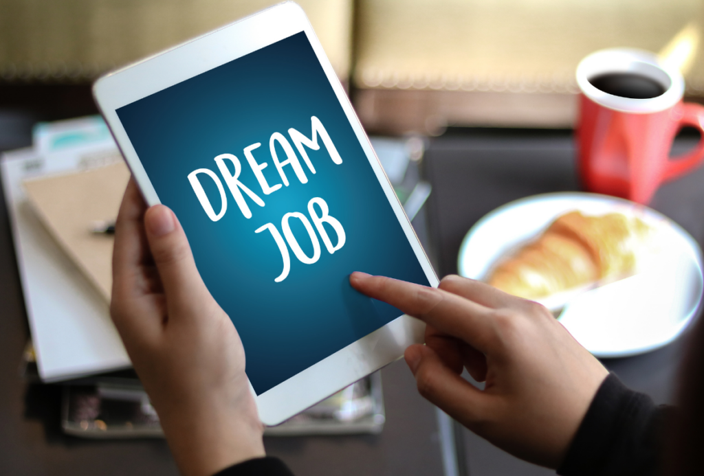 Dłonie trzymające tablet. Na tablecie wyświetla się na niebieskim tle biały napis "dream job". W tle widać biurko z notatnikami, kubkiem i talerzem z rogalikiem.