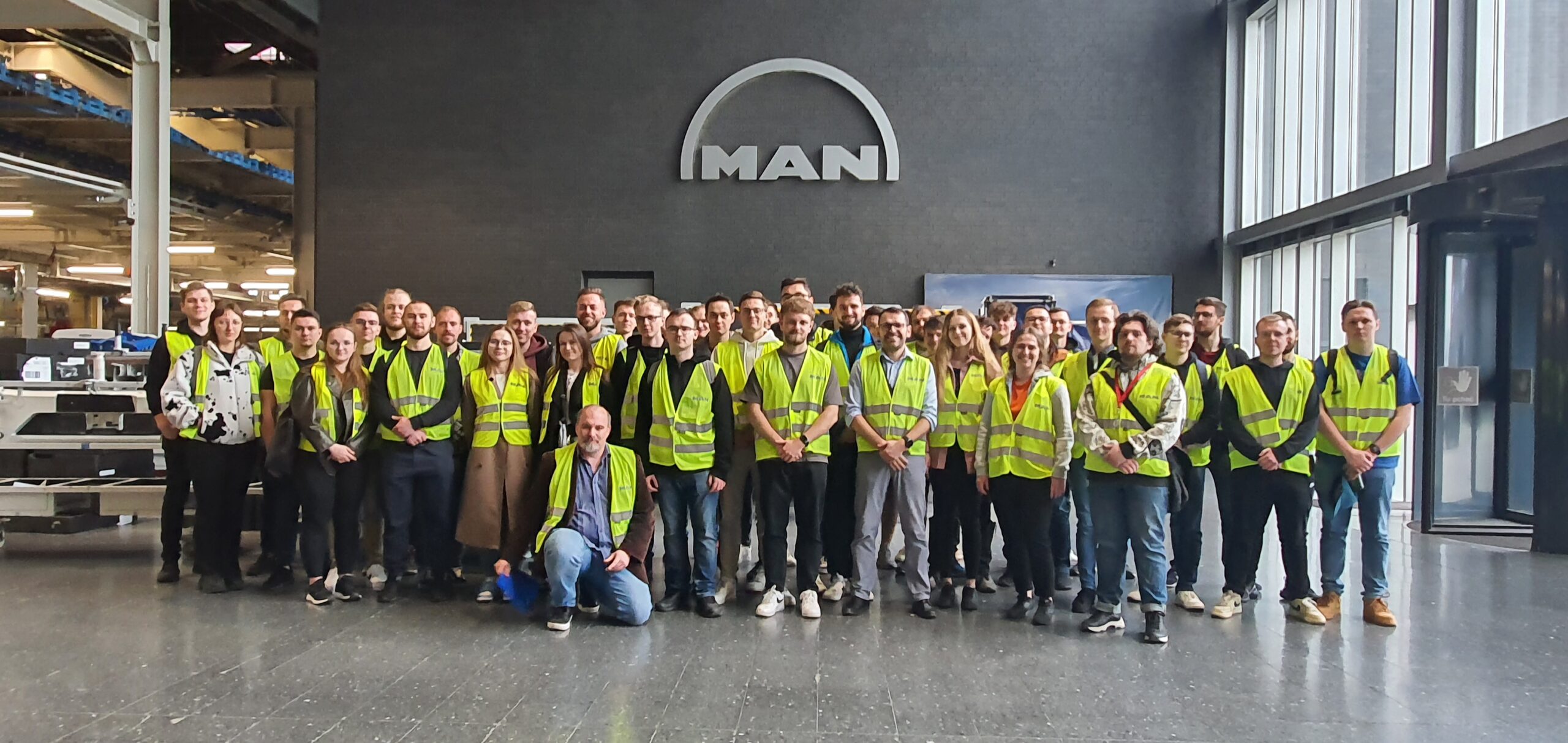 Grupa studentów w odblaskowych kamizelkach ochronnych pozuje do wspólnego zdjęcia. Za mini na szarej ścianie duże logo firmy MAN.