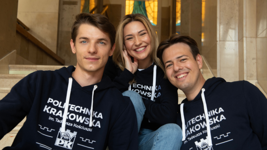 Troje studentów, dwóch mężczyzn i kobieta. Cała trójka ma na sobie granatowe logo z białym logo Politechniki Krakowskiej. Uśmiechają się.