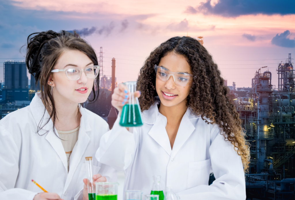 Dwie młode kobiety w białych fartuchach laboratoryjnych i okularach ochronnych pracują w laboratorium. Jedna z nich trzyma kolbę z zielonym płynem i uśmiecha się, druga notuje coś ołówkiem. W tle widać panoramę zakładów przemysłowych przy zachodzie słońca