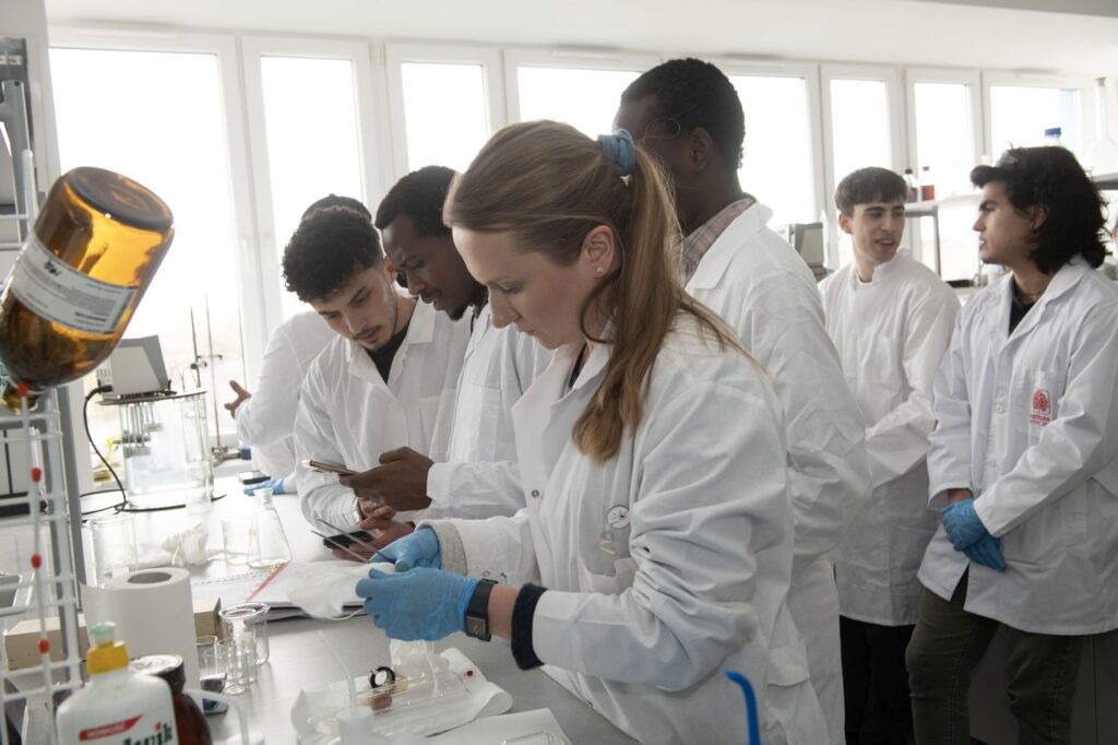 Grupa studentów w białych fartuchach laboratoryjnych pracuje w nowoczesnym laboratorium. Na pierwszym planie studentka w niebieskich rękawiczkach wykonuje precyzyjne czynności przy stanowisku, obok niej dwóch studentów sprawdza coś na telefonie. W tle widać innych studentów rozmawiających ze sobą. Na stole znajdują się kolby, pipety, ręczniki papierowe i sprzęt laboratoryjny.