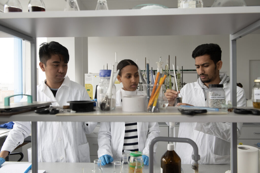 Troje studentów w białych fartuchach laboratoryjnych pracuje w laboratorium. Stoją przy stanowisku pełnym szkła laboratoryjnego, probówek, narzędzi i reagentów. Jeden ze studentów trzyma fiolkę i uważnie ją ogląda, podczas gdy pozostali obserwują jego działania. W tle widać sprzęt laboratoryjny oraz półki z odczynnikami.