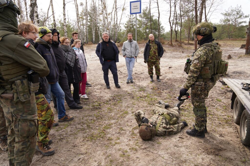 Grupa osób w ubraniach cywilnych stoi wokół żołnierza Wojska Polskiego. U jego stóp, na ziemi, leży drugi żołnierz.