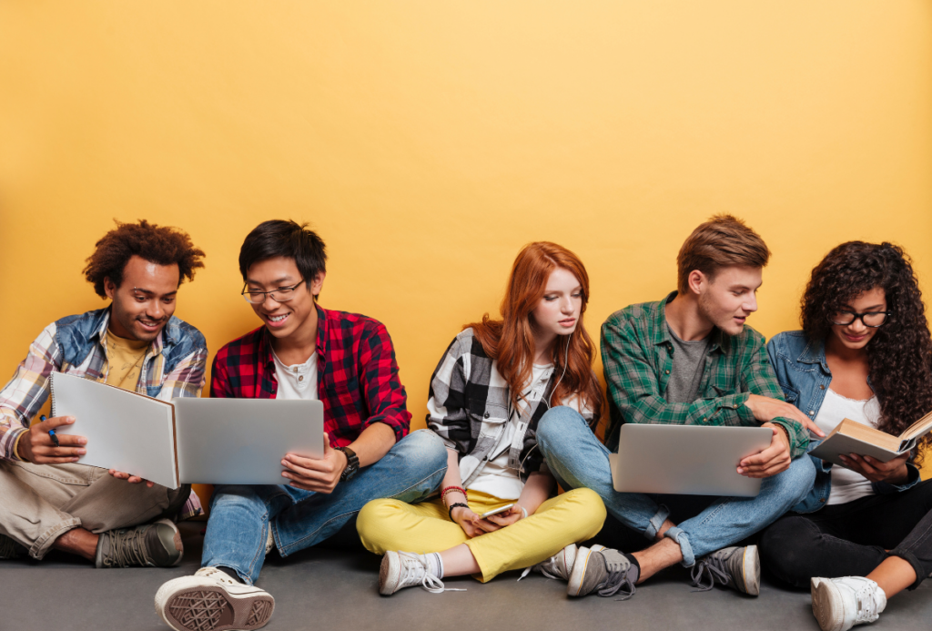Pięcioro młodych ludzi w kolorowych ubraniach, trzech chłopaków i dwie dziewczyny siedzących na podłodze. Patrzą na ekrany trzech laptopów i w ksiązkę. W tle pomarańczowa ściana