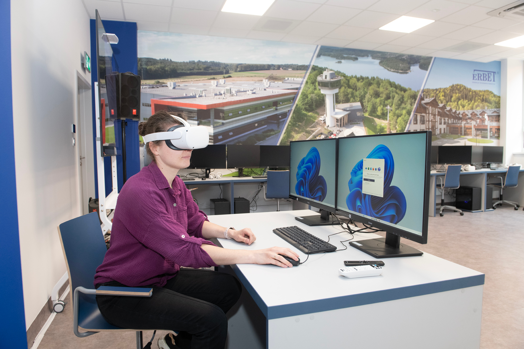 Młoda kobieta w fioletowej bluzce siedzi przy biurku z dwoma monitorami. Na oczach ma google VR. W tle widać nowocześnie wyposażoną salę komputerową.