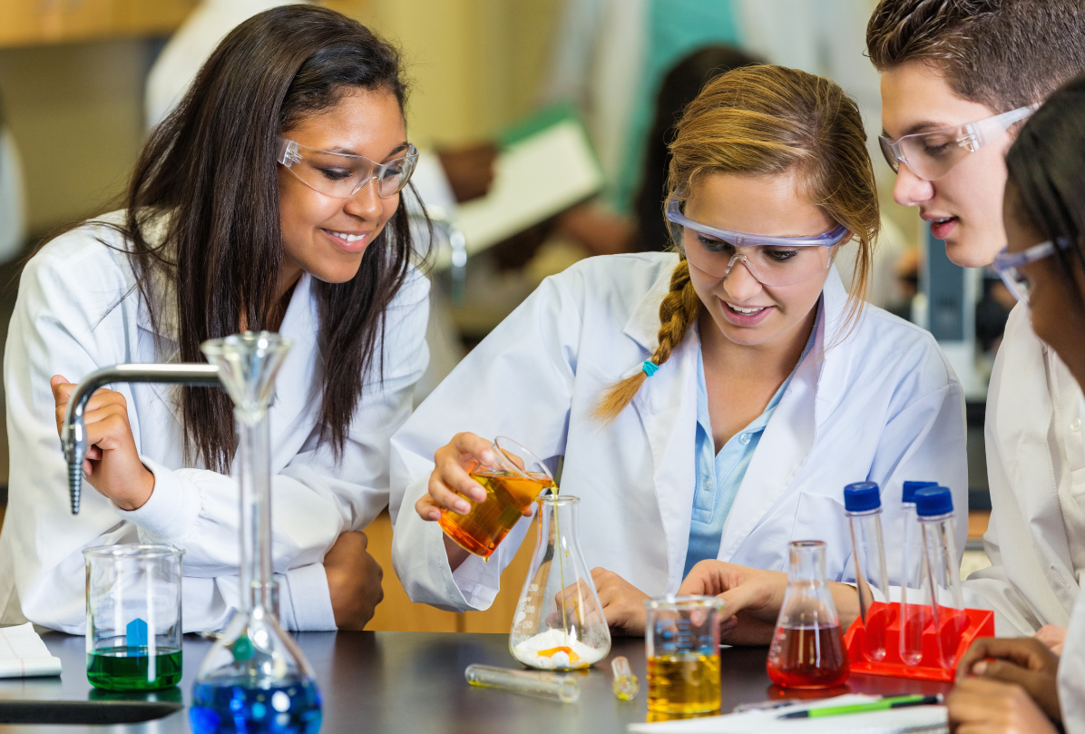 Troje studentów w białych fartuchach prowadzi eksperyment chemiczny. Wszyscy się uśmiechają i chyba dobrze bawią.